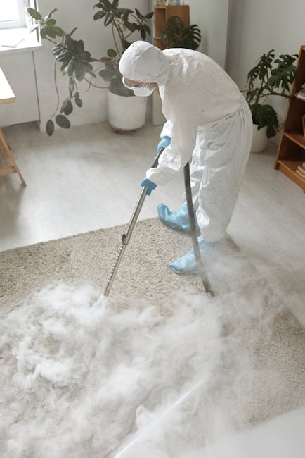 A professional cleaner dressed in a full white protective suit, including gloves, a face mask, and shoe covers, is performing deep cleaning on a light-colored carpet in a bright, modern room. The clea