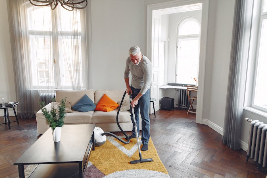 A person standing barefoot on a beige textured rug in a room with wooden flooring, using a red and black upright vacuum cleaner for surface cleaning. The individual is dressed in black pants and a mat